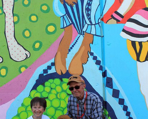 A couple pose with their dog under the golden retriever wearing a ball gown on the Kellogg Park Dog Field mural by Artworks