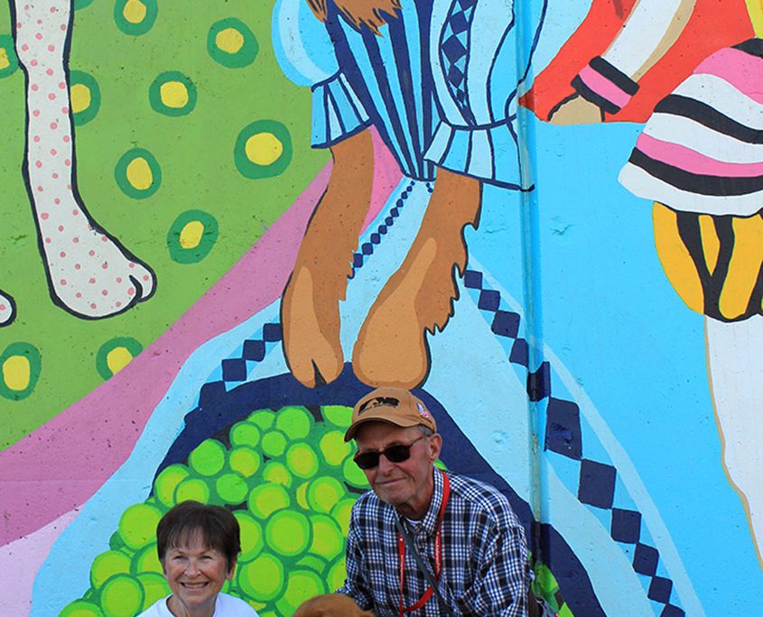 A couple pose with their dog under the golden retriever wearing a ball gown on the Kellogg Park Dog Field mural by Artworks