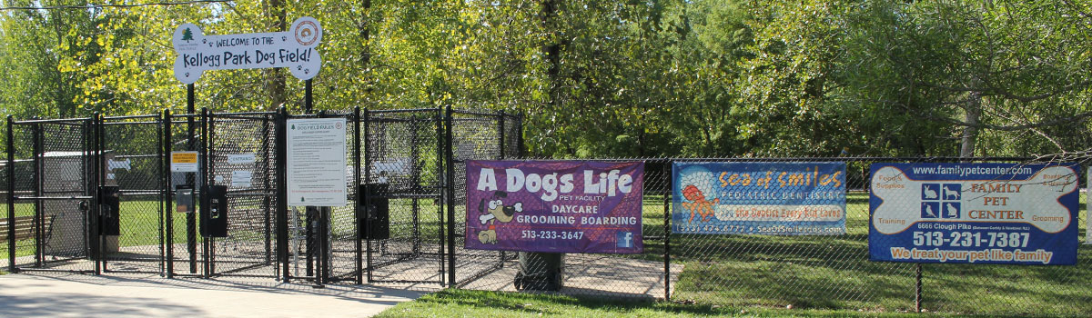 The gated entrance to Kellogg Park Dog Field. Above the gate is a dog-bone shaped sign reading "Welcome to the Kellogg Park Dog Field!" in a playful font. The park rules are posted near the entrance, and the fence is adorned with several colorful advertising banners.